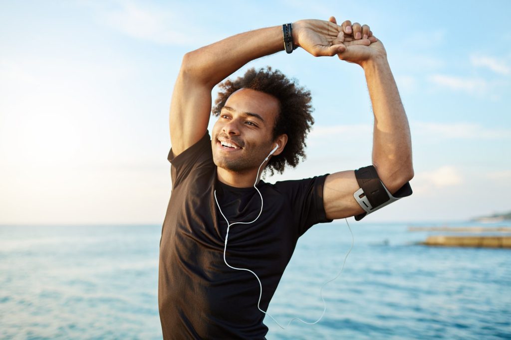 Person wearing a black shirt and earphones stretching arms overhead near a body of water.