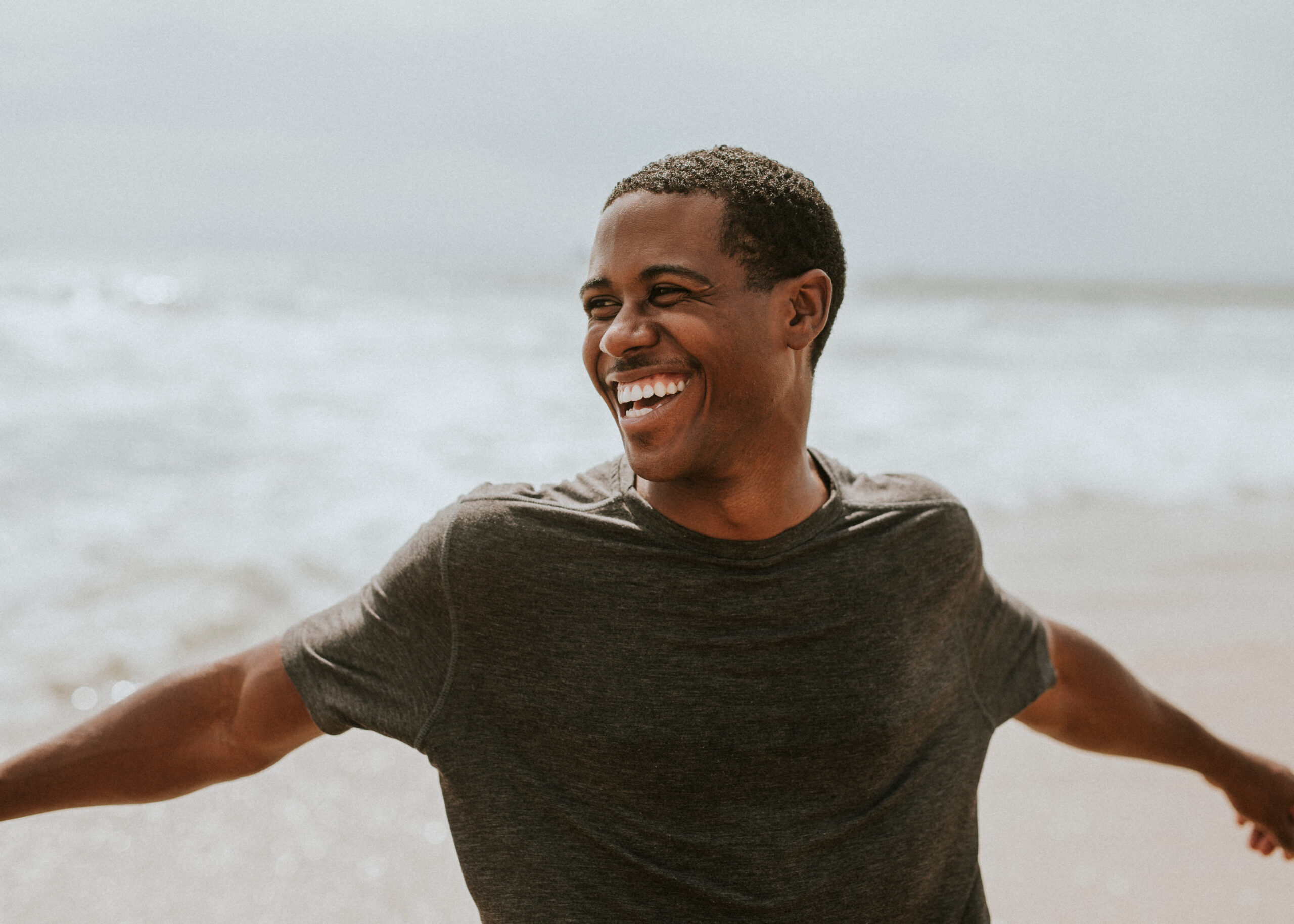 A man in a dark T-shirt stands on a beach with arms outstretched while smiling toward the side. Image Description: