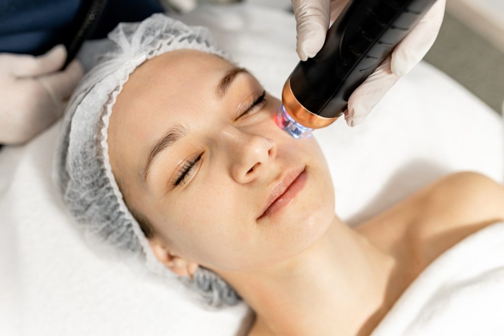 A person lies on a treatment bed while a gloved practitioner applies a handheld device with light to the cheek area.